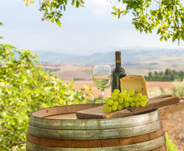 A wine bottle, a wine glass, a block of cheese and a bunch of grapes on a wine cask